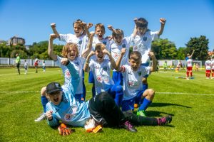 Eine Gruppe kleiner Kinder in Fu&szlig;balltrikots feiert begeistert auf einer Wiese, reckt triumphierend die Arme in die H&ouml;he, w&auml;hrend ein Kind vor der Gruppe liegt. Der Himmel ist klar und blau.