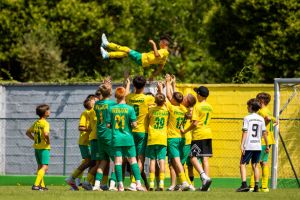 Eine Gruppe von Kindern in gelben und gr&uuml;nen Fu&szlig;balltrikots feiert, indem sie einen Mitspieler in die Luft werfen, w&auml;hrend ein paar andere Kinder auf einem sonnigen Feld daneben zuschauen.