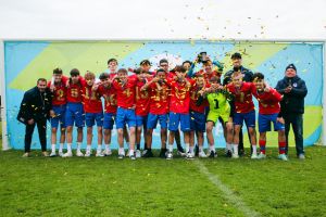A football team in red and blue kits celebrates on the pitch, standing in front of a colourful wall as confetti falls. Two adults stand at the ends, and all are smiling or cheering.