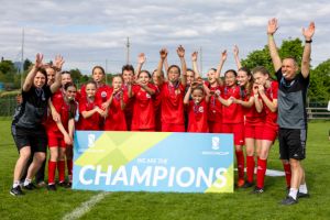 A joyful girls' football team in red kits celebrates behind a large Champions banner on a grass pitch, raising their arms and smiling at the camera. Two coaches stand on either side, also cheering.