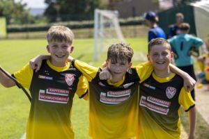 Three smiling boys in yellow and black football kits stand together with arms round each other on a grassy pitch, with a goal and other children in the background.